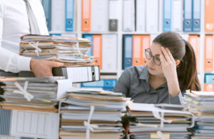 Young stressed secretary in the office overwhelmed by work and desk full of files, her boss is bringing more paperwork to her