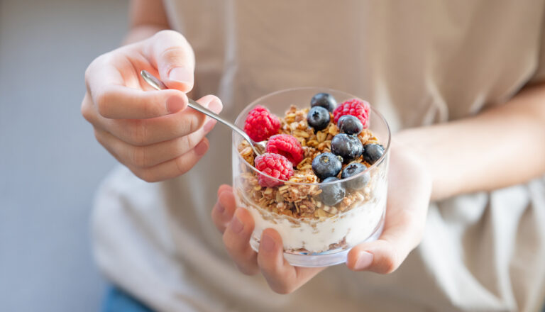 Granola with greek yogurt, raspberries and blueberries in a glass in hands of a woman in a light t-shirt close up. Dairy breakfast concept, oat muesli in the morning light.
