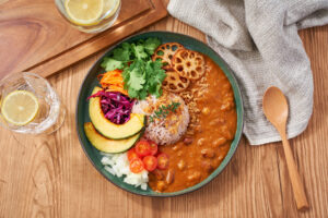 A vibrant and healthy curry plate featuring mixed grain rice, hearty bean curry, and a colorful array of vegetables like pumpkin, lotus root, cherry tomatoes, and carrots. Garnished with fresh coriander and served on a flat plate, this vegetarian dish offers a perfect balance of nutrition and flavor.