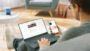 Young Man at Home is Using Laptop Computer, Srufing the Net, Watching News, while Holding Smartphone with Social Media Interface. He's Sitting On a Couch in His Cozy Living Room. Over Shoulder Shot