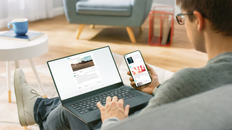 Young Man at Home is Using Laptop Computer, Srufing the Net, Watching News, while Holding Smartphone with Social Media Interface. He's Sitting On a Couch in His Cozy Living Room. Over Shoulder Shot
