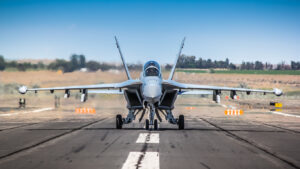 The  Growler is a specialized electronic warfare version of the U.S. Navy's two-seat F/A-18F Super Hornet jet fighter. The image depicts a full, frontal shot of the jet aircraft on the taxi way with engines running.   Hot exhaust gases are blurring the air behind the jet.  Electronic counter measures pods can be seen underneath each wing.  The sky is a crystal clear blue.