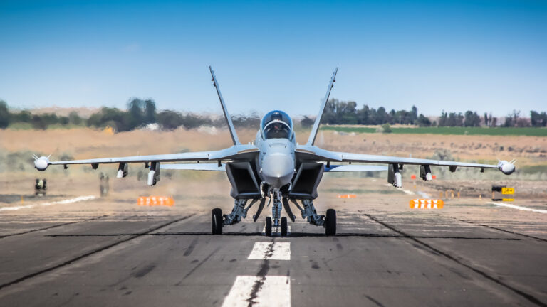 The  Growler is a specialized electronic warfare version of the U.S. Navy's two-seat F/A-18F Super Hornet jet fighter. The image depicts a full, frontal shot of the jet aircraft on the taxi way with engines running.   Hot exhaust gases are blurring the air behind the jet.  Electronic counter measures pods can be seen underneath each wing.  The sky is a crystal clear blue.